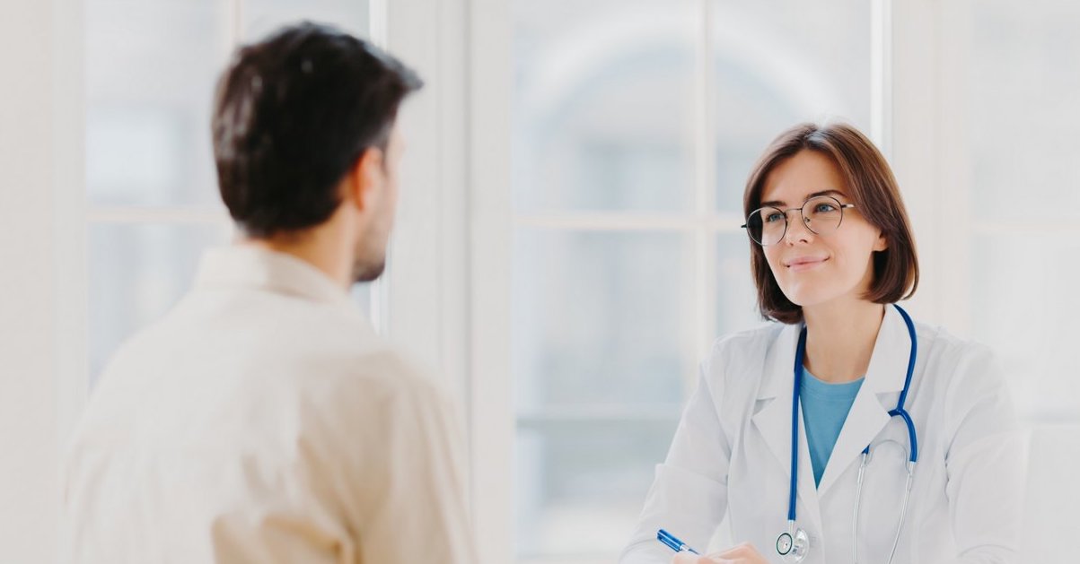 Doctor and patient discuss something, sit at table in clinic. Female cardiologist in eyewear gives medical consultation diagnostic, advice for man how to cure disease, pose in hospital room.